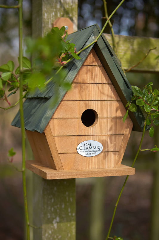 Wooden birdhouse with green roof attached to a tree, featuring the brand 'Tom Chambers'.