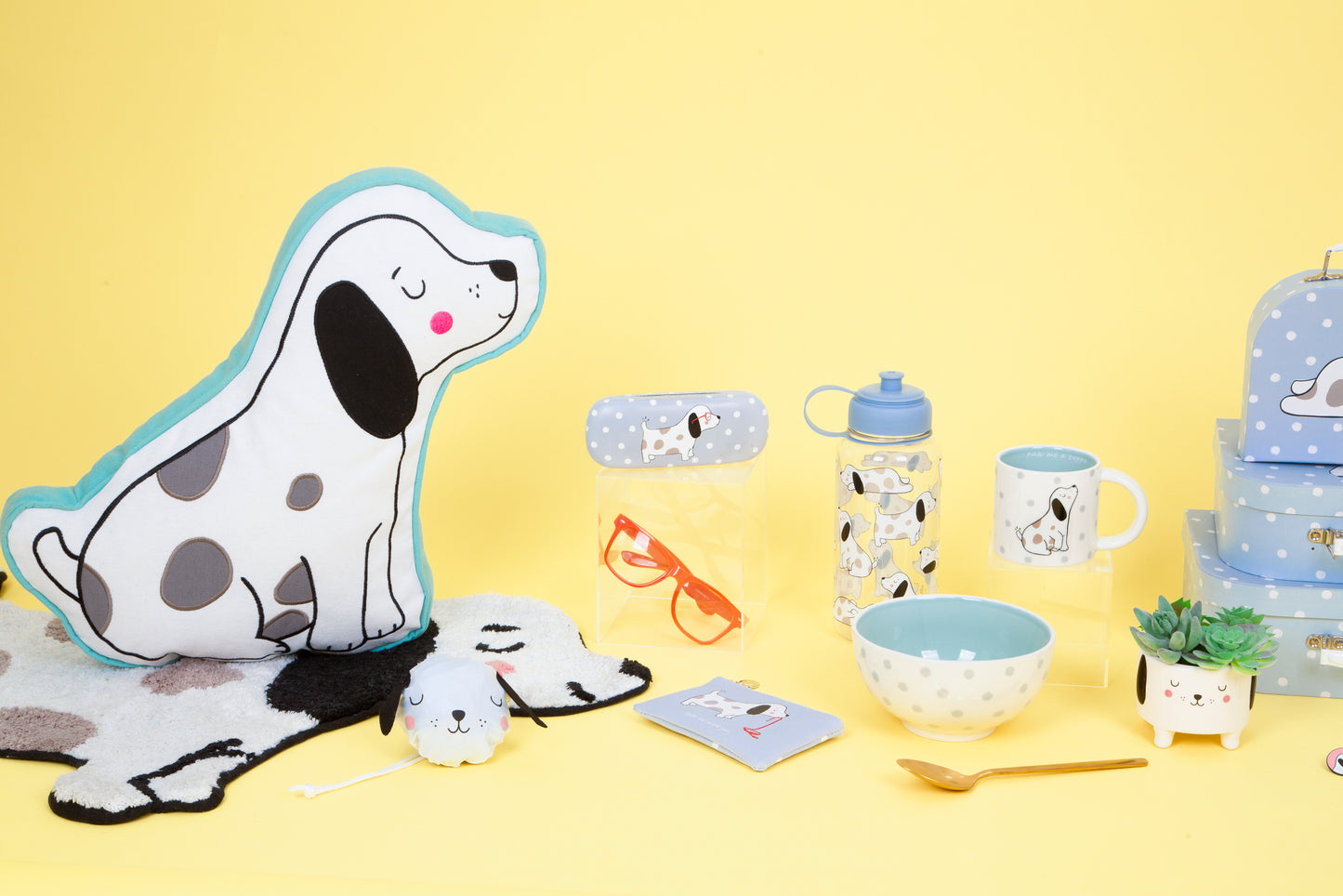 Collection of children's items including a dog-shaped pillow, cups, bowls, and backpack on a yellow background.