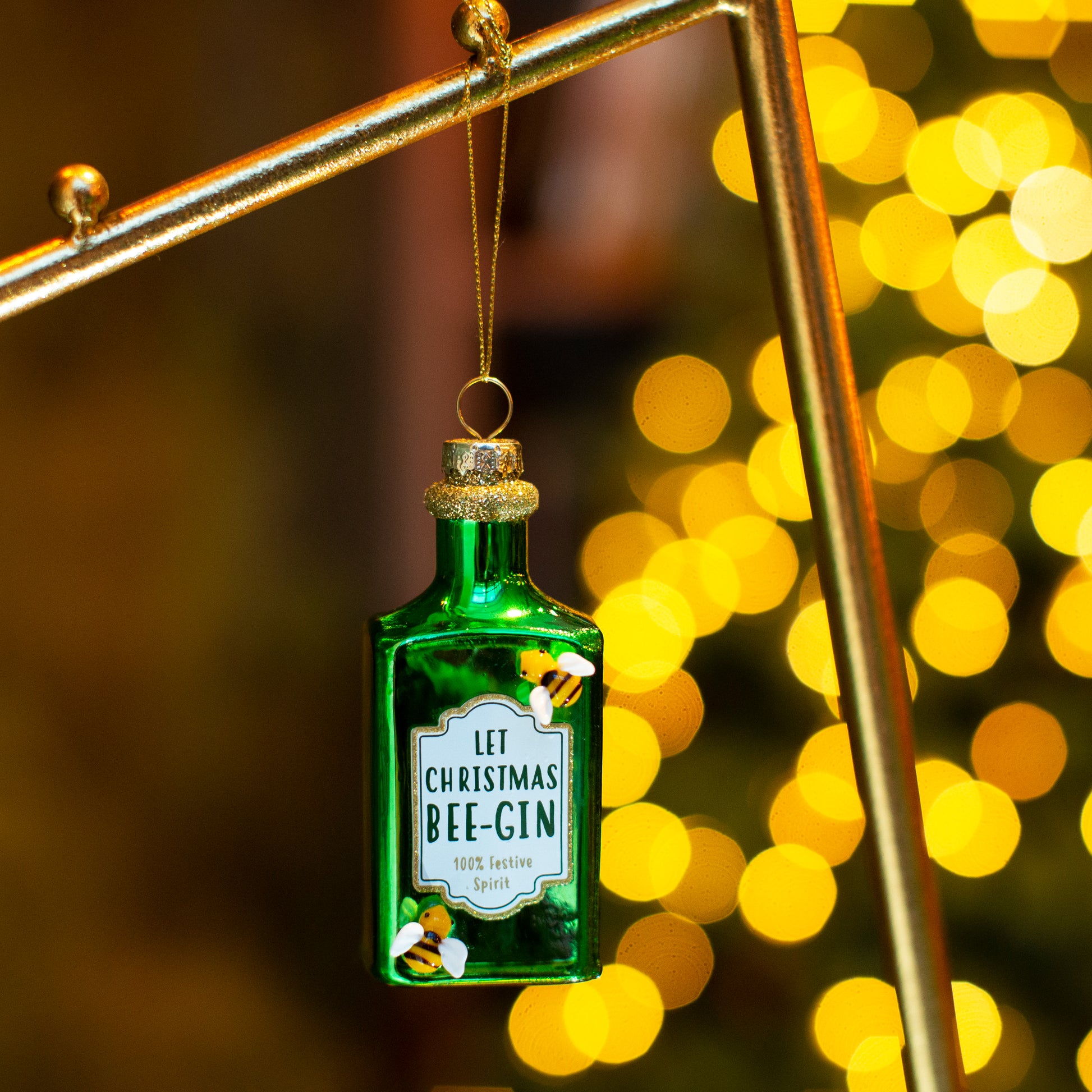 Green bottle-shaped ornament with 'Let Christmas Bee-Gin' text against a blurred festive background.