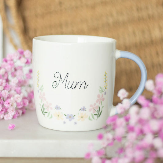 White mug with 'Mum' and floral designs, surrounded by pink flowers on a wooden surface.
