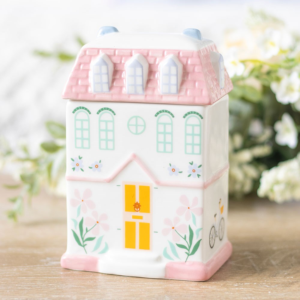 Ceramic house-shaped container with pink roof and yellow door on a wooden surface with blurred greenery in the background
