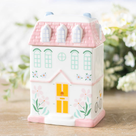 Ceramic house-shaped container with pink roof and yellow door on a wooden surface with blurred greenery in the background