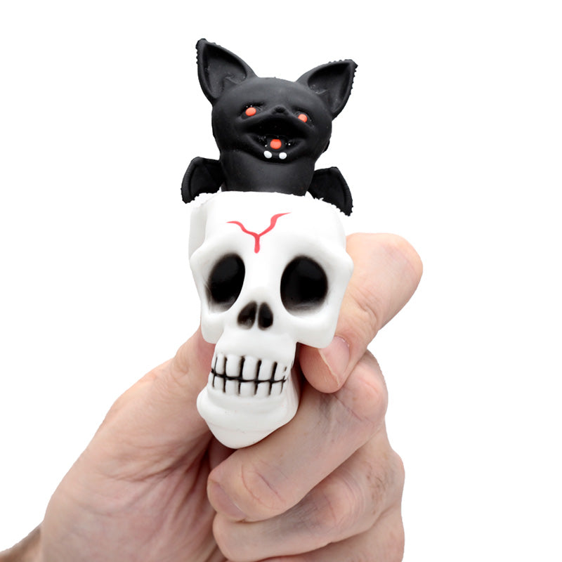 Black bat figurine on top of a white skull with red eyes held by a hand against a white background
