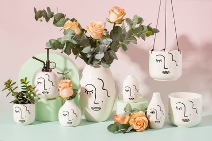 Hanging planter with white pot featuring a face design with other similar pots and vases on a pink and green background