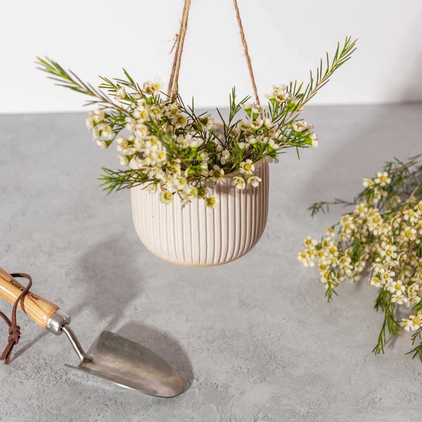 White hanging planter with greenery on a gray surface with a trowel.