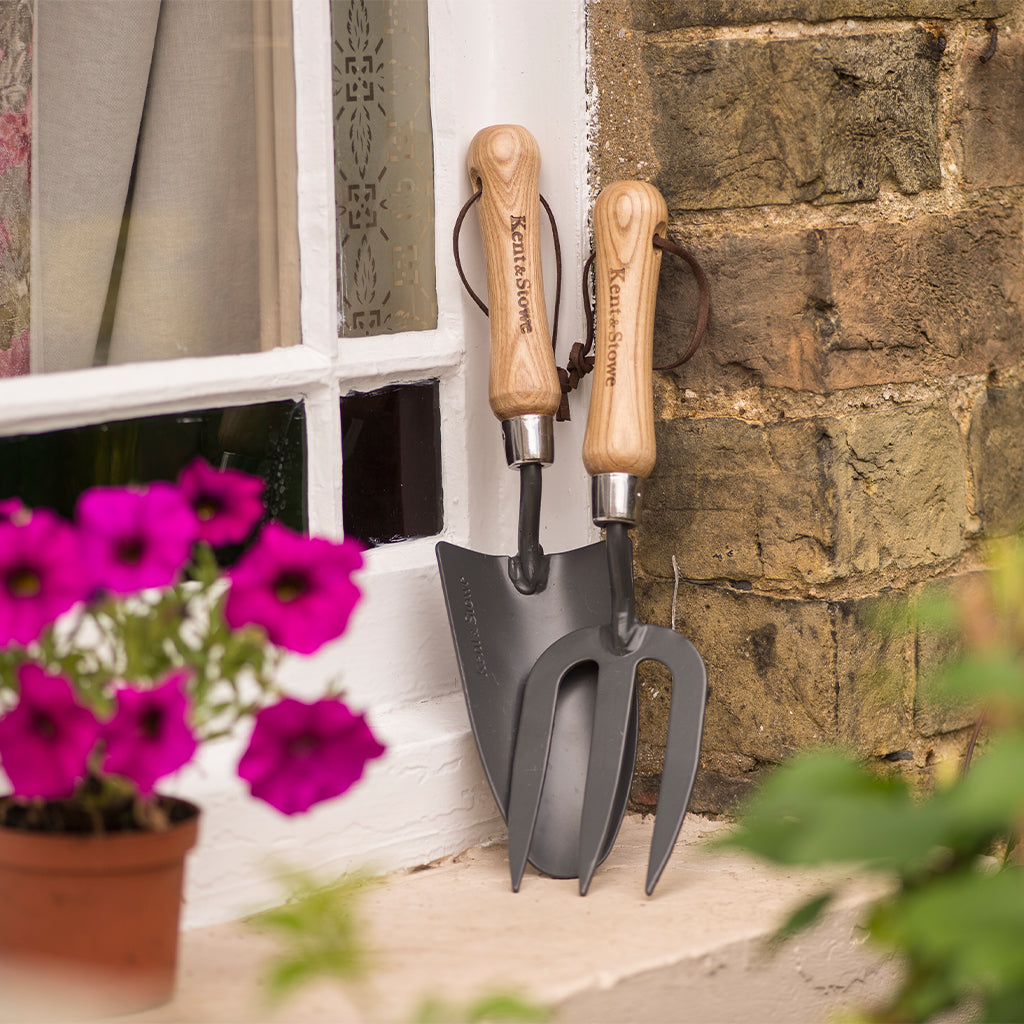 Garden trowel and fork with wooden handles against a stone wall and window.