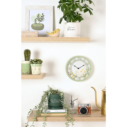 Decorative wall clock with plants and books on a shelf against a white wall.
