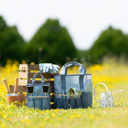 Gardening tools including a blue tool chest, trowel, gloves, and watering can on grass with blurred trees in the background.