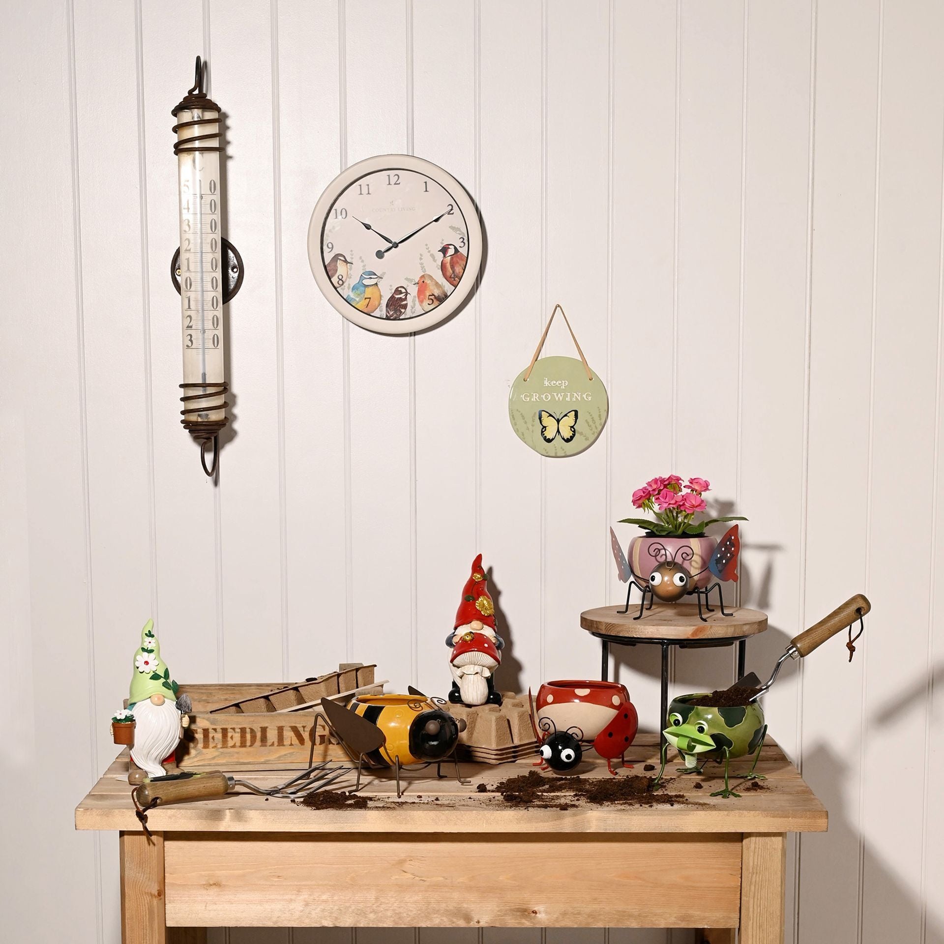 Decorative items including a clock, thermometer, and small plants on a wooden table against a white paneled wall.