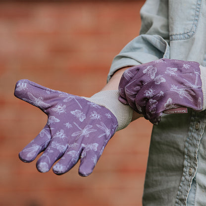 Purple gardening gloves with white patterns worn by a person against a blurred brick wall background.