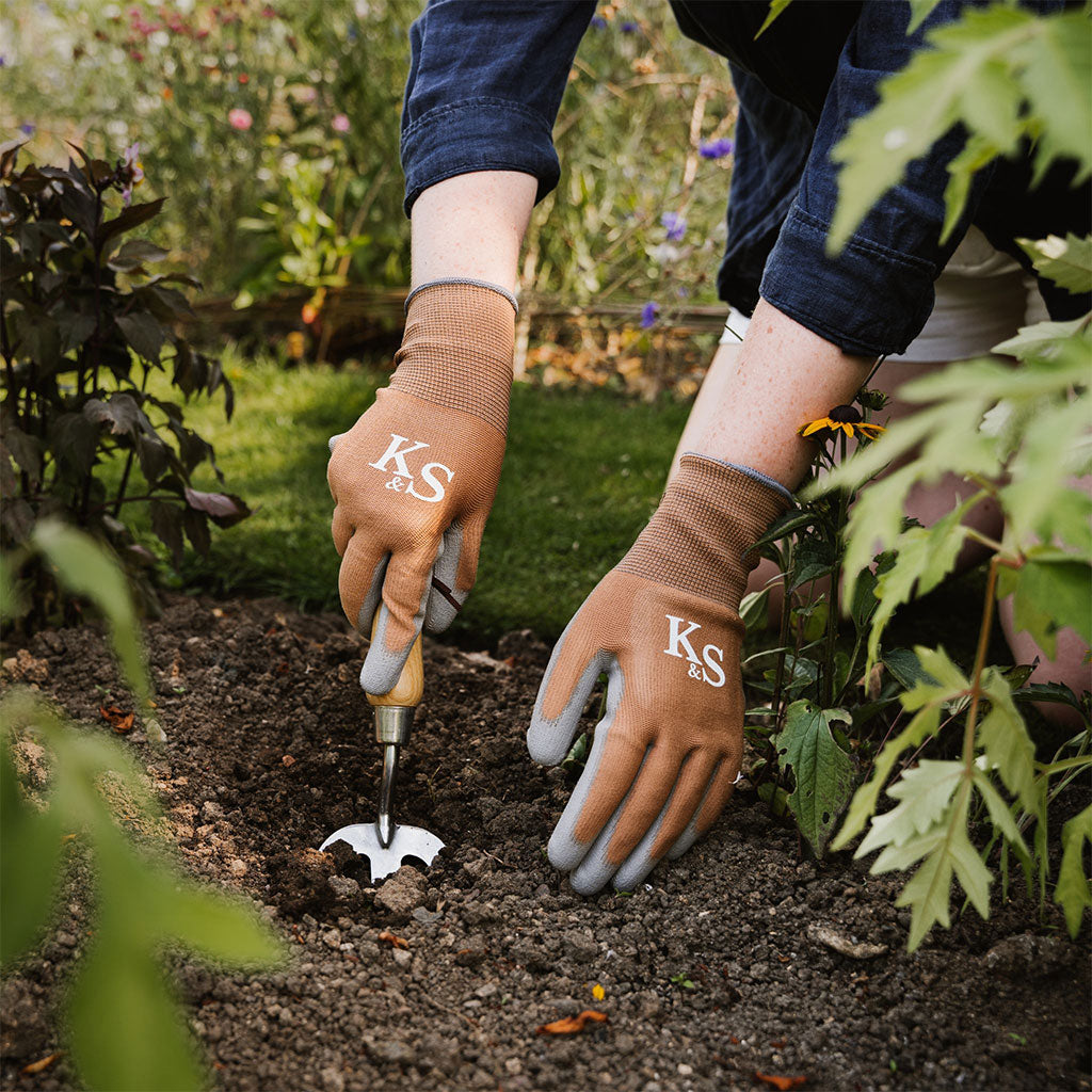 Person gardening with brown gloves labeled 'KS' in a garden setting.