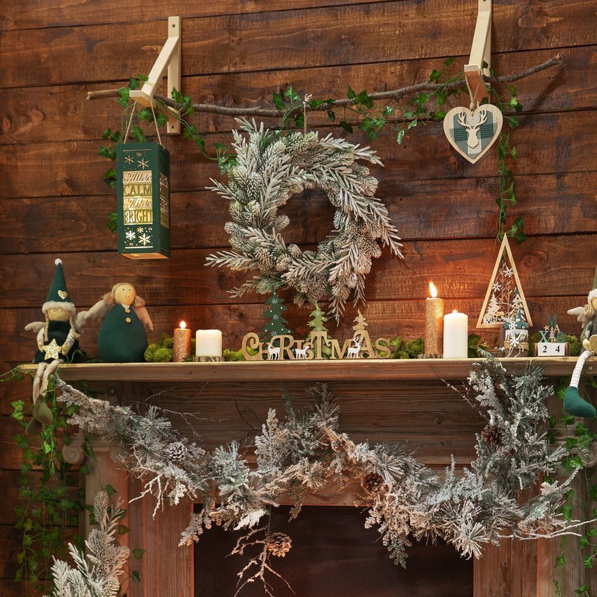 Decorative Christmas mantel with wreaths, candles, and ornaments against a wooden wall.