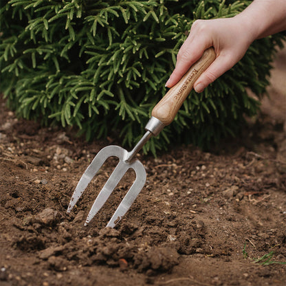 Hand holding a gardening fork in soil with green plants in the background