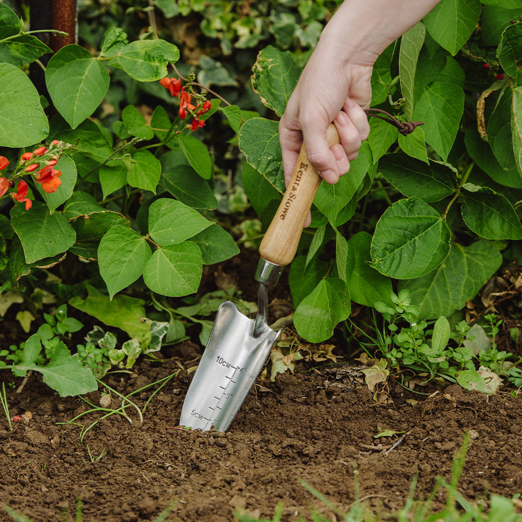 Hand using a small gardening trowel in soil with plants and flowers in the background