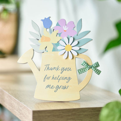 Decorative card shaped like a watering can with flowers and text on a wooden surface.