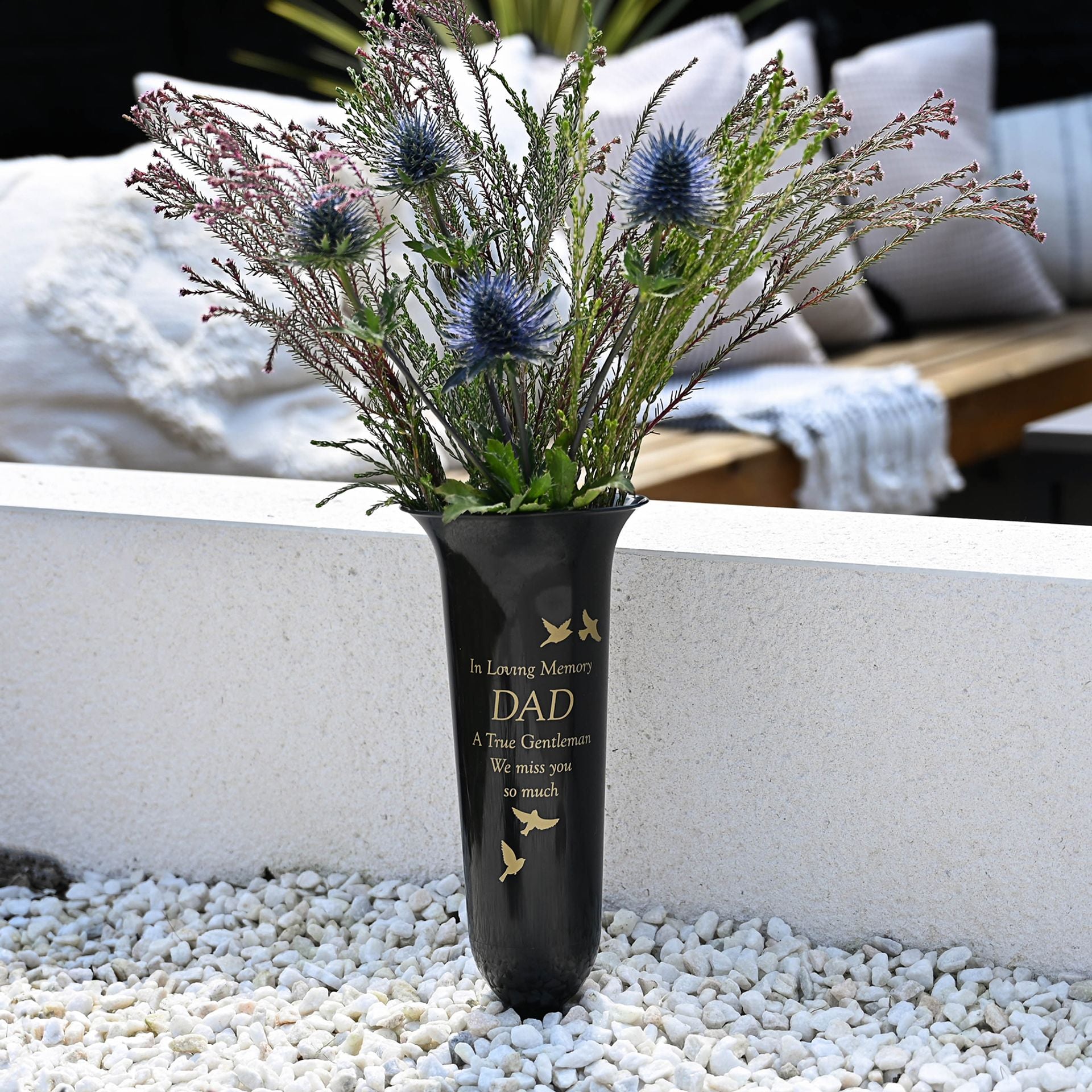 Black vase with floral arrangement on pebbles, text 'In Loving Memory DAD A True Gentleman We Miss You So Much' against a blurred outdoor background.