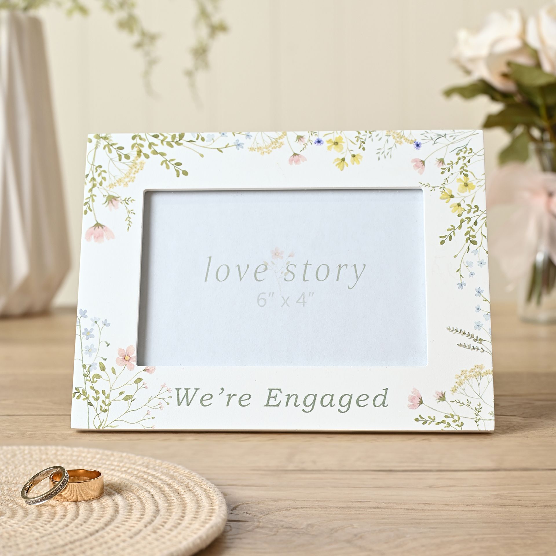 Floral photo frame with 'love story' and 'We're Engaged' text on a wooden surface.