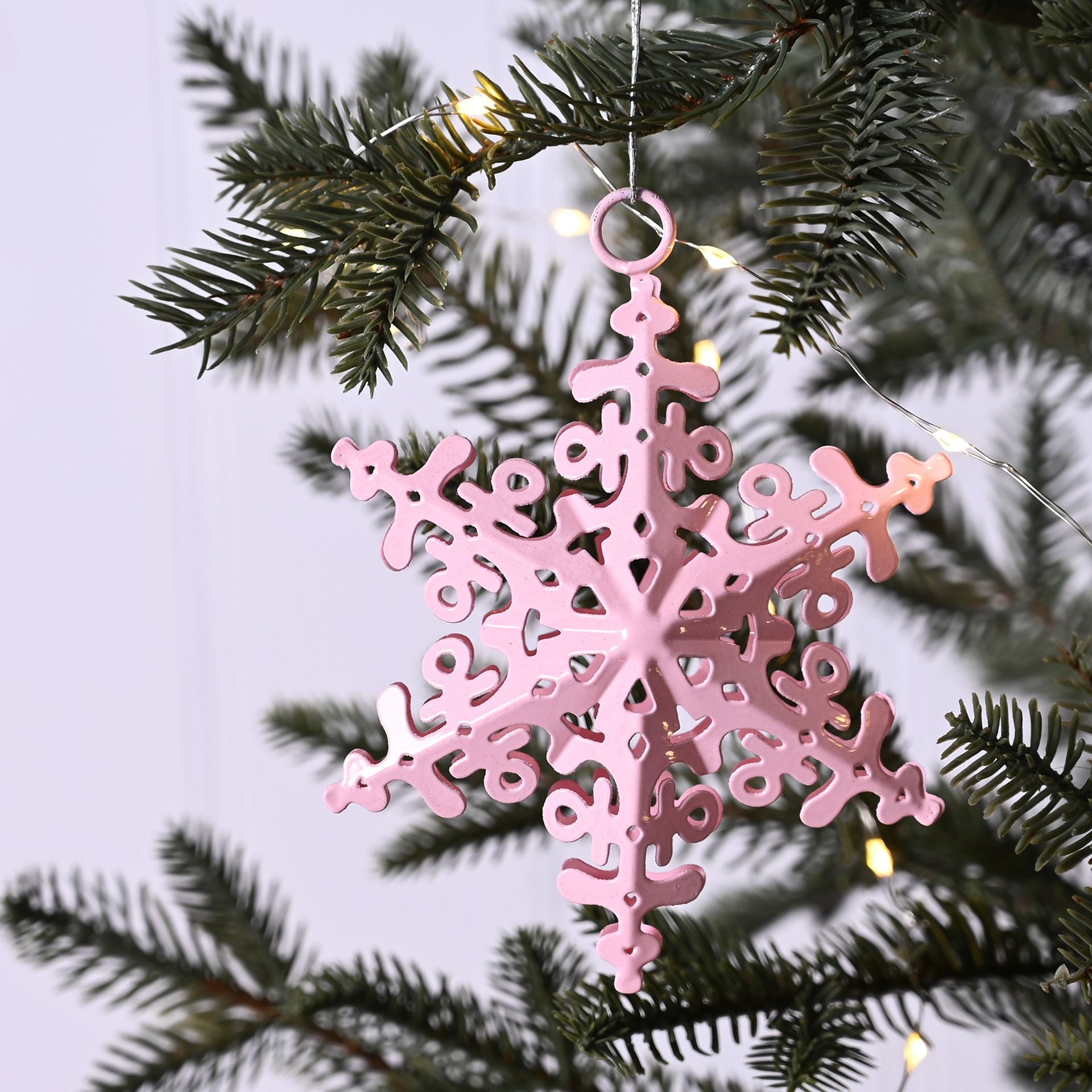 Pink snowflake ornament hanging on a Christmas tree branch with lights.