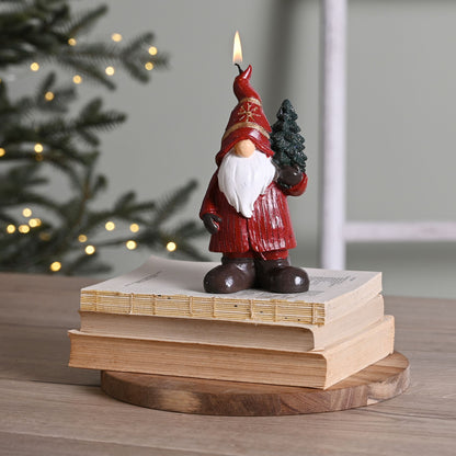 Decorative candle shaped like a gnome holding a small tree, standing on books with a blurred Christmas tree in the background.