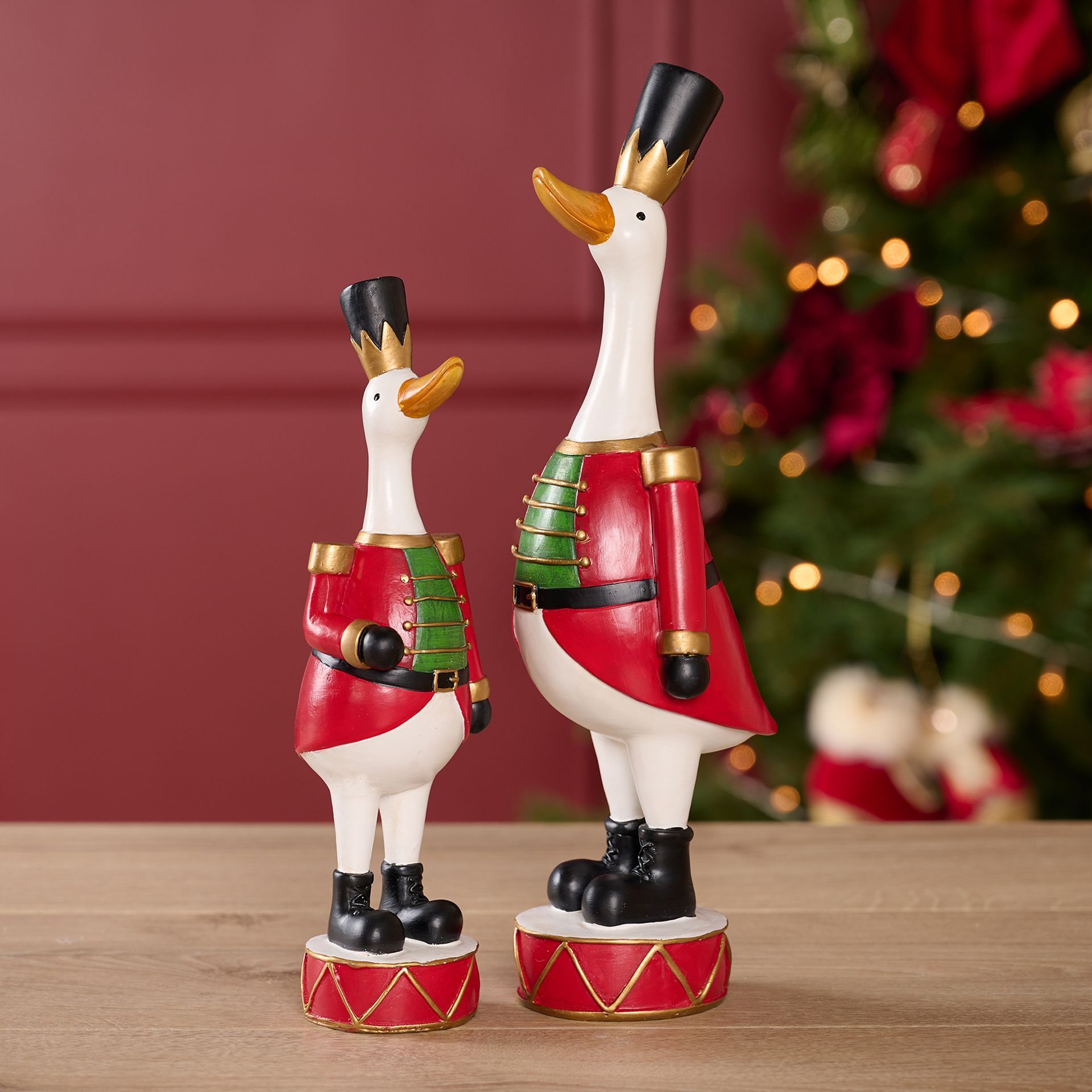 Two decorative goose figurines in festive costumes on a wooden surface with a Christmas tree in the background.